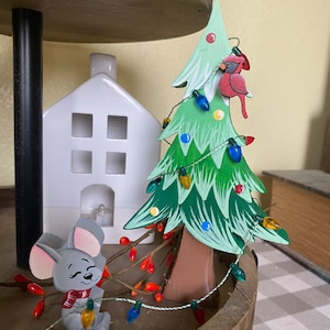 Puede incluir: Un árbol de Navidad de madera con detalles pintados en verde y azul, decorado con luces de colores y un adorno de cardenal rojo. Una figurita de ratón gris con una bufanda roja sostiene una cadena de luces.