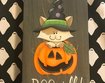 May include: A wooden sign featuring a painted image of a cat wearing a witch's hat and holding a jack-o'-lantern. The sign reads "BOO, y'all!" in white paint.