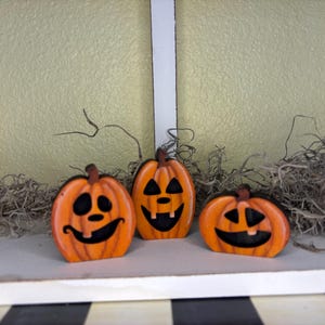 Può includere: Tre zucche di legno arancioni con facce nere e gambi marroni. Le zucche sono disposte in fila, con muschio secco e uno sfondo giallo chiaro. Una decorazione festosa di Halloween.