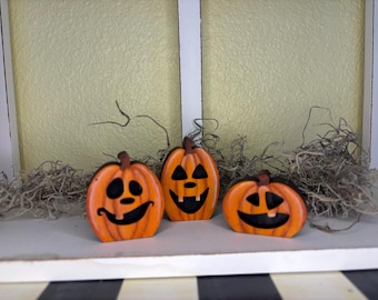 May include: Three orange wooden jack-o'-lanterns with black facial features and brown stems. The pumpkins are arranged in a row, with dried moss and a light yellow background. The pumpkins are a festive Halloween decoration.