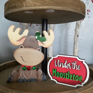 May include: A wooden tiered tray with Christmas decorations. The bottom tier features a smiling moose with antlers, a mistletoe sprig, and a sign that reads "Under the Moostletoe". The top tier has a wooden figure with a red hat.
