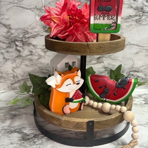 May include: A two-tier wooden tiered tray with a black metal stand. The top tier has a red flower, a wooden popsicle with the text "hello summer" and an ant, and the bottom tier has a wooden fox holding a watermelon, a watermelon slice with ants, and a wooden bead garland with a jute tassel.