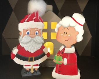 May include: A wooden Santa Claus and Mrs. Claus figurine set. Santa is wearing a red suit with white trim and a white beard. Mrs. Claus is wearing a red dress with white trim and a white hat. They are both holding a small green gift.