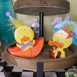 May include: Two wooden duck figurines, one holding an orange umbrella and the other wearing a blue umbrella hat. Both ducks are painted yellow with black eyes and orange beaks. The duck with the umbrella hat has a red and white bow.
