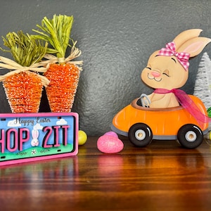 May include: An Easter-themed display featuring a wooden bunny driving an orange car, a license plate that says "HOP 2 IT", and two carrot decorations. The bunny wears a pink bow and scarf. Decorative Easter eggs are scattered around.