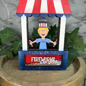 May include: A wooden figurine of a person wearing a red, white, and blue hat and shirt holding fireworks. The figurine is in front of a red, white, and blue striped awning with the word "FIREWORKS" written on the front.