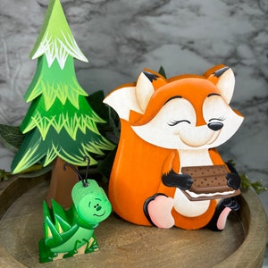 May include: A wooden figurine set featuring a fox holding a chocolate sandwich cookie, a green grasshopper, and a green pine tree. The figurines are painted with bright colors and have a whimsical style.