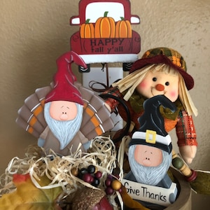 May include: A Thanksgiving themed tiered tray featuring a red truck with the text "Happy Fall y'all" and three gnomes. One gnome is a turkey, one is a scarecrow, and the other is a pilgrim. The tray is decorated with autumn leaves, acorns, and berries.