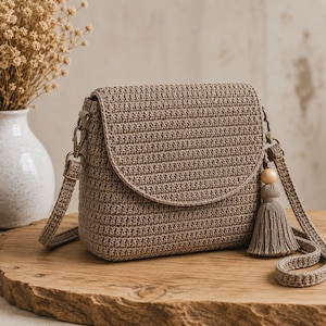 Può includere: Una borsa a tracolla all'uncinetto color tortora con patta arrotondata, dotata di tracolla in coordinato fissata con ganci in metallo color bronzo. Una nappa decorativa con una perlina in legno chiaro pende dal lato della borsa.
