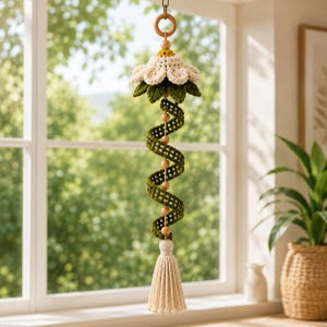 May include: A hanging crochet decoration featuring a white flower with a yellow center, suspended from a wooden ring. A green spiral vine with small wooden beads runs down the center, ending in a cream-colored tassel at the bottom.