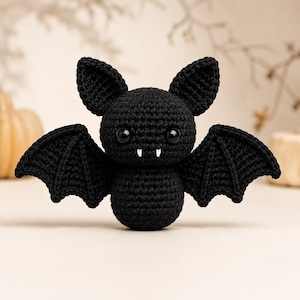 May include: A black crocheted bat plush toy featuring large pointed ears, two glossy black safety eyes, and two small white fangs protruding from the mouth. The bat has outstretched wings with visible stitching detail and a round body.