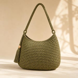 May include: An olive green crochet shoulder bag featuring a textured stitch pattern, a single integrated handle, and a decorative tassel accent attached to a wooden bead on the side. The bag has a rounded silhouette and a consistent yarn construction.