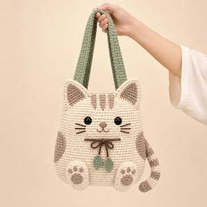May include: A crochet tote bag shaped like a cat with cream-colored yarn, brown tabby stripes, and two long sage green handles. The cat features black button eyes, embroidered whiskers, a small brown nose, and a decorative bow with two green leaves.