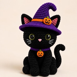 May include: A black crocheted cat figurine wearing a purple witch hat with an orange band and a small pumpkin accent. The cat has large green eyes, a pink nose, white stitched whiskers, and a purple collar featuring a hanging orange pumpkin charm.