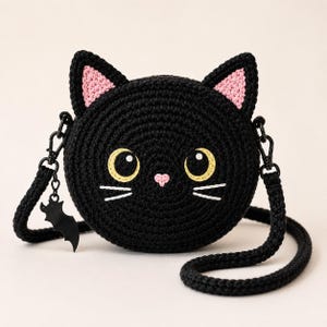 May include: A round black crochet crossbody bag shaped like a cat face with pointed ears, pink inner ear details, large yellow eyes, a small pink nose, and white embroidered whiskers. A black bat-shaped charm hangs from the side strap attachment.