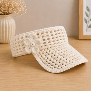 May include: A cream-colored crochet visor featuring an open-weave grid pattern and a single, dimensional crocheted flower accent attached to the side. The visor has a structured brim and a band that wraps around the head, crafted from yarn.