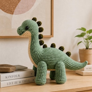 Può includere: Un dinosauro giocattolo all'uncinetto di colore verde chiaro con ventre color crema e punte verde scuro lungo la schiena. Il peluche presenta una testa arrotondata, grandi occhi neri, quattro zampe robuste e una lunga coda in filato.