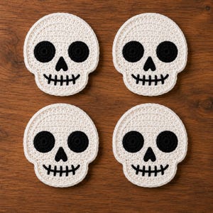 Könnte beinhalten: Ein Set aus vier runden Häkeluntersetzern, die jeweils ein weißes Totenkopfdesign mit großen schwarzen runden Augenhöhlen, einer kleinen schwarzen dreieckigen Nase und einer breiten, schwarz gestickten Mundlinie in einem Raster zeigen.