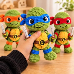 May include: Three handmade crochet turtle dolls with large black eyes. Each doll wears a colored eye mask, matching wristbands, and a yellow chest plate with a brown belt featuring a circular emblem with the letter L. Each holds two small gray swords.