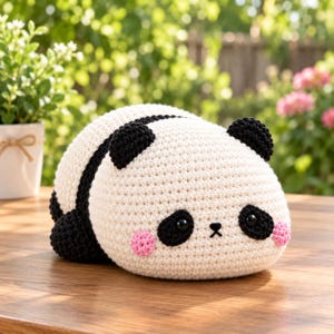 May include: A crochet panda plush toy featuring a rounded white body with black ears, black eye patches, and small black limbs. The face includes two black plastic eyes, a small black embroidered nose, and two round pink cheeks on the sides.