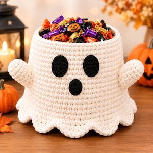 Può includere: Un cestino a forma di fantasma lavorato all'uncinetto color crema con un bordo inferiore smerlato, due piccole braccia e tre toppe ovali nere che formano un volto. L'interno cavo è riempito con varie caramelle colorate incartate, inclusi disegni di zucche arancioni.