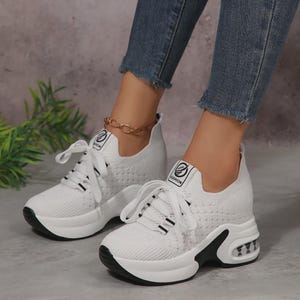 Könnte beinhalten: Ein Paar weiße Schnür-Sneaker mit einer dicken Plateausohle mit schwarzen Akzenten und einem transparenten Luftpolster an der Ferse. Das Obermaterial besteht aus strukturiertem Strickstoff mit einem kleinen rechteckigen Etikett mit der Aufschrift FASHION.
