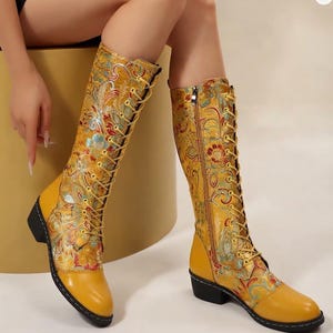 May include: A pair of knee-high boots featuring a vibrant yellow base with intricate floral embroidery in shades of red, blue, and gold. These boots include a front lace-up closure with yellow laces, a side zip, and a low, solid black heel.