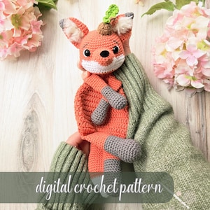 May include: A handmade crochet fox doll in shades of orange, white, and gray, with a green leaf detail on its head. The doll is held against a background of pink flowers and the text "digital crochet pattern".