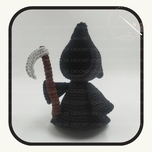 May include: A crocheted Grim Reaper doll with a black hooded robe and a gray scythe with a brown handle. The doll is handmade and has a whimsical design. The background is a light cream color.