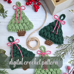 May include: Crocheted Christmas tree ornaments in shades of green, with brown trunks and red and white striped bows. The ornaments are arranged on a white surface with decorative greenery and berries. The text "digital crochet pattern" is visible.