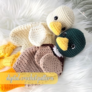Puede incluir: Juego de juguetes de pato de ganchillo en crema, marrón, amarillo y verde oscuro. El juego incluye un cuerpo de pato, dos cabezas de pato y una pancarta amarilla con el texto "digital crochet pattern". Los juguetes están colocados sobre una superficie blanca y esponjosa.