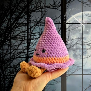 May include: A plush crochet toy shaped like a pointed hat with a purple top, an orange band, and tan feet. The toy features small black safety eyes and a stitched smile. The texture is soft and fuzzy, crafted from thick yarn in a rounded, conical form.