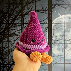 May include: A plush, cone-shaped crochet toy in deep magenta and light purple with a wide brim. It features two small black safety eyes, a tiny stitched mouth, pink blush marks, and two tan, rounded feet dangling from the bottom edge.