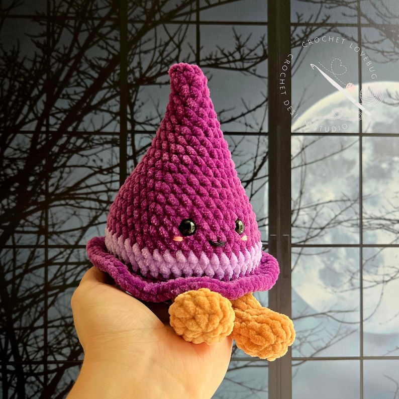 May include: A plush, cone-shaped crochet toy in deep magenta and light purple with a wide brim. It features two small black safety eyes, a tiny stitched mouth, pink blush marks, and two tan, rounded feet dangling from the bottom edge.
