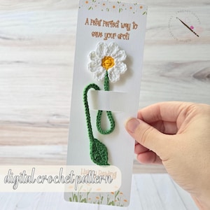 Könnte beinhalten: Häkel-Lesezeichen in Form einer Gänseblume. Die weiße Blume mit gelber Mitte sitzt auf einem grünen Stiel und Blatt. Das Lesezeichen ist an einer weißen Karte mit dem Text "A Polar Perfect way to save your spot" und "digital crochet pattern" befestigt.