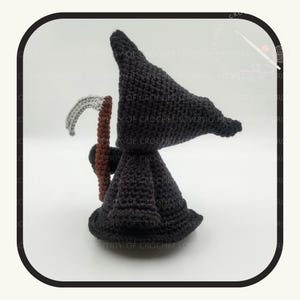 May include: A crocheted figure of the Grim Reaper, featuring a black hooded cloak and a scythe. The scythe has a gray blade and a brown handle. The figure is handmade and stands on a white surface.