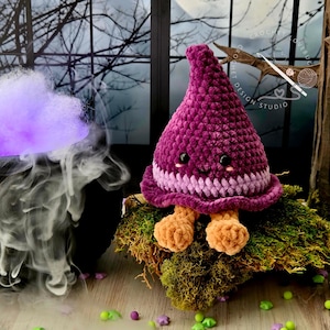 May include: A plush, crocheted witch hat character with a deep purple pointed top, a lighter purple band, and two small tan feet. The character features black safety eyes, a small embroidered smile, and pink blush marks on its face.