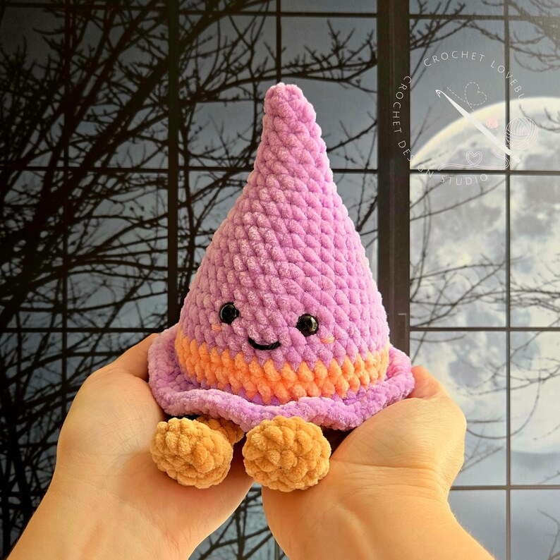 May include: A plush, cone-shaped crochet toy featuring a light purple textured body with a wide orange band near the base. The toy has two small black bead eyes, a stitched black smile, and two small tan feet dangling from the bottom edge.