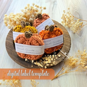 May include: A wooden bowl holds several handmade crochet facial scrubbies in the shape of pumpkins. The scrubbies are orange and yellow, with brown stems. Each scrubby has a white label that reads "Facial Scrubbies". Dried flowers surround the bowl.