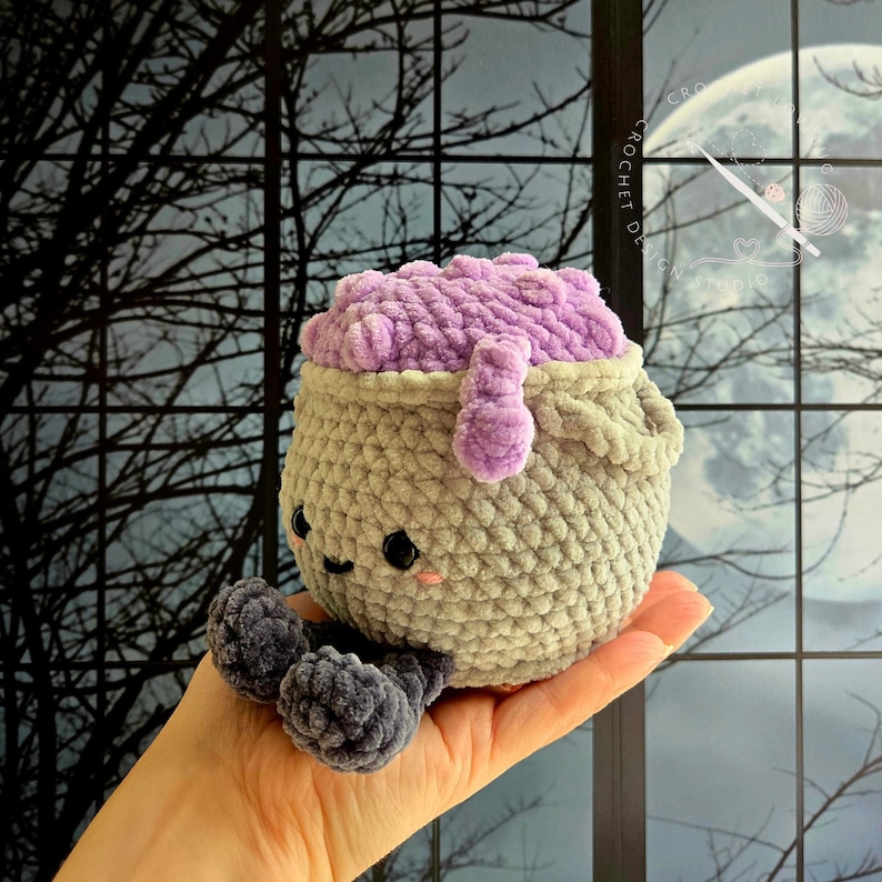 Op de afbeelding: Een gehaakt ketel-knuffeltje met een lichtgrijs lichaam en een textuurrijke paarse bubbelende bovenkant. De ketel heeft twee kleine zwarte ogen, een geborduurde glimlach en twee donkergrijze bungelende benen. Het oppervlak is zacht.
