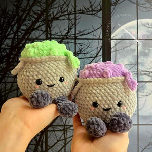 Op de afbeelding: Twee ronde, gehaakte knuffels in de vorm van ketels. De ene heeft een lichtgroene bovenkant en de andere een paarse, beide met grijze lichamen, donkergrijze voetjes, zwarte veiligheidsoogjes en kleine roze geborduurde wangetjes.