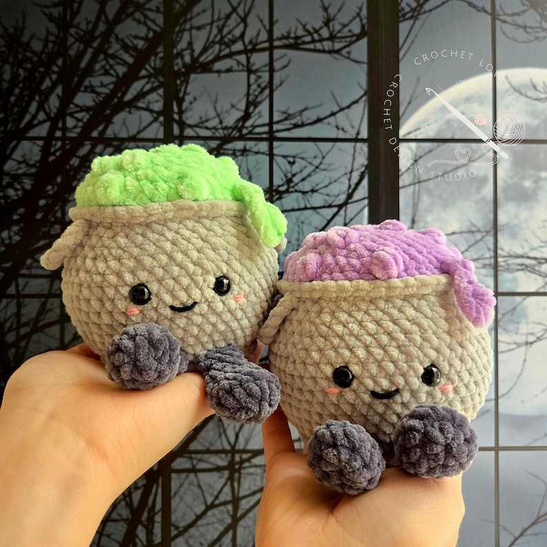 Op de afbeelding: Twee ronde, gehaakte knuffels in de vorm van ketels. De ene heeft een lichtgroene bovenkant en de andere een paarse, beide met grijze lichamen, donkergrijze voetjes, zwarte veiligheidsoogjes en kleine roze geborduurde wangetjes.