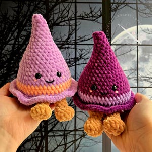 May include: Two plush crochet figures shaped like pointed hats with small round feet. The left figure is light purple with an orange band, and the right figure is dark magenta with a light purple band. Both have black eyes and stitched smiles.