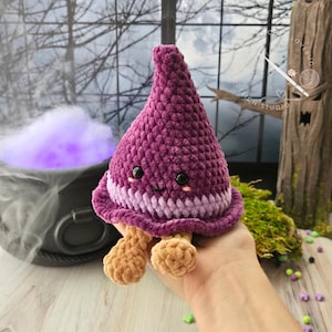 May include: A plush, cone-shaped crochet toy in deep purple with a lighter purple band at the base. It features two small black safety eyes, a stitched black smile, two tiny pink blush marks, and two small, light tan, rounded feet dangling below.