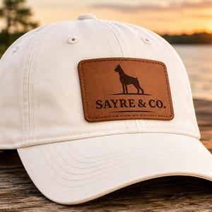 May include: A cream-colored baseball cap featuring a rectangular brown leather patch on the front. The patch displays a black silhouette of a Doberman dog above the text SAYRE & CO. in a bold, dark font, with decorative horizontal lines.