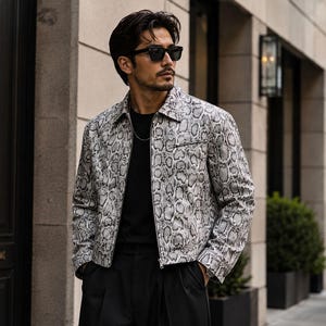 Könnte beinhalten: Eine langärmelige Jacke mit grau-schwarzem Schlangenledermuster, spitzem Kragen und durchgehendem Frontreißverschluss. Die Jacke wird über einem schlichten schwarzen T-Shirt getragen und mit einer schwarzen Hose kombiniert. Dazu eine schwarze Sonnenbrille.