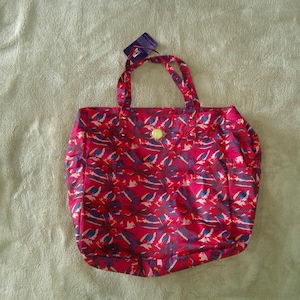 May include: A red tote bag featuring a repeating abstract pattern in shades of blue and white. The bag has two top handles and a small, circular, light-colored emblem centered on the front. A dark purple rectangular tag is attached to one handle.