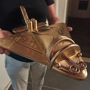 Op de afbeelding: Een goudkleurig, driedimensionaal model van een ruimtevaartuig met een driehoekige vleugelstructuur, een cilindrische neussectie en decoratieve spiraalpatronen op het oppervlak. Het object heeft een metallic afwerking met zichtbare laagstructuren.