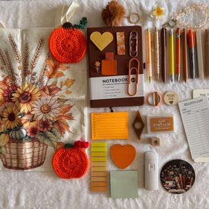 May include: A collection of stationery items including a canvas tote bag with a pumpkin and sunflower print, a brown A5 notebook, two crocheted pumpkin coasters, various orange and yellow pens, sticky notes, paper clips, and a reading list card.