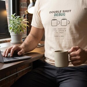 May include: A beige t-shirt featuring the text DOUBLE SHOT DEBUG, two coffee mug icons, and the code lines: brew --double-shot, trace: caffeine.ok, fix: ship(), and the brand name CADENCE & CREMA printed in dark brown and green ink.
